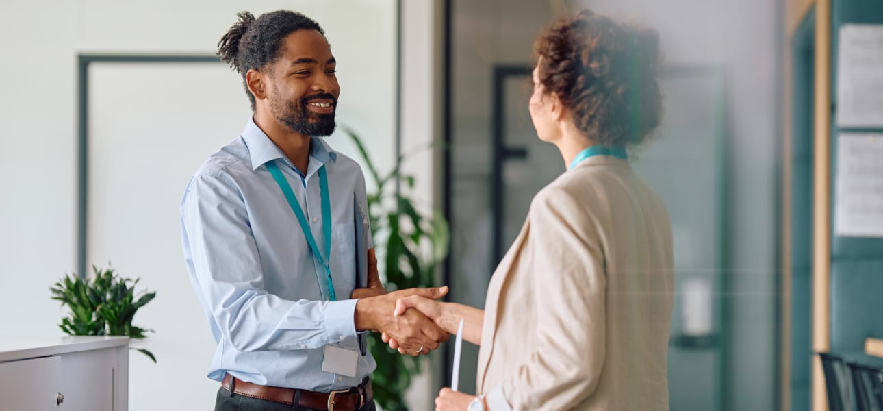 Man in a blue button down shirt shakes hands with a woman in a business office.