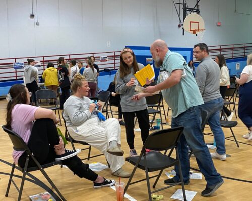 Adults stand or sit around metal folding chairs in a gymnasium.