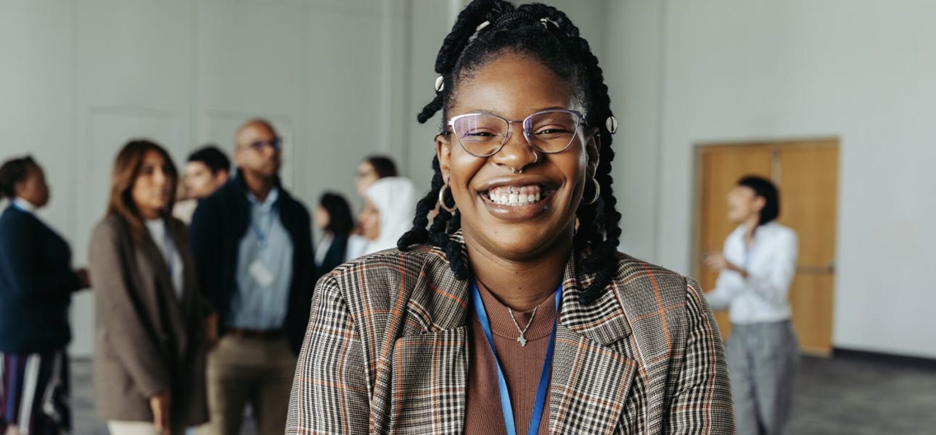 Woman wearing glasses and a plaid blazer smiles; other business colleagues talk in the background.