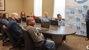 Diverse group of adults sit around a long table, looking towards a woman giving a presentation.