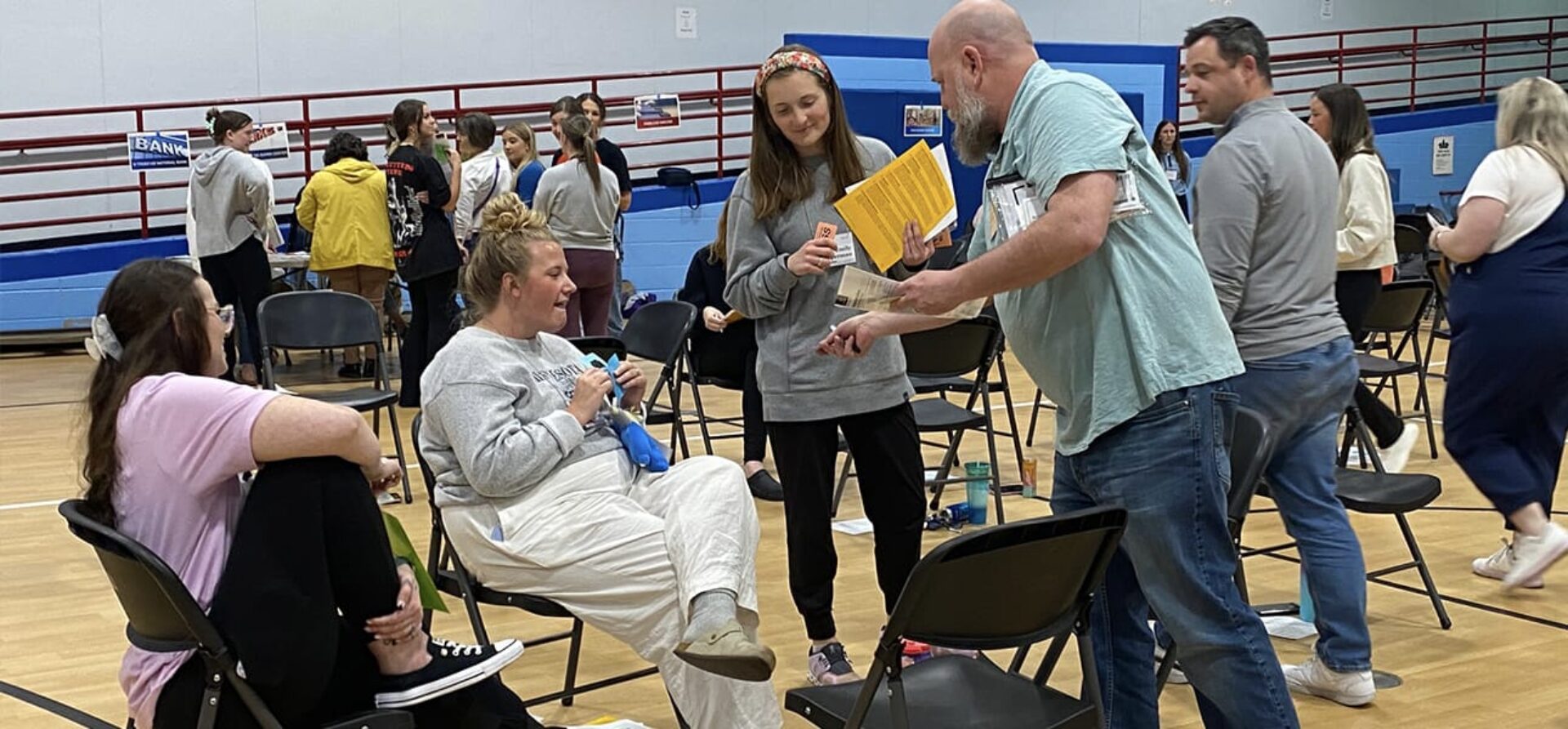 Adults stand or sit around metal folding chairs in a gymnasium.