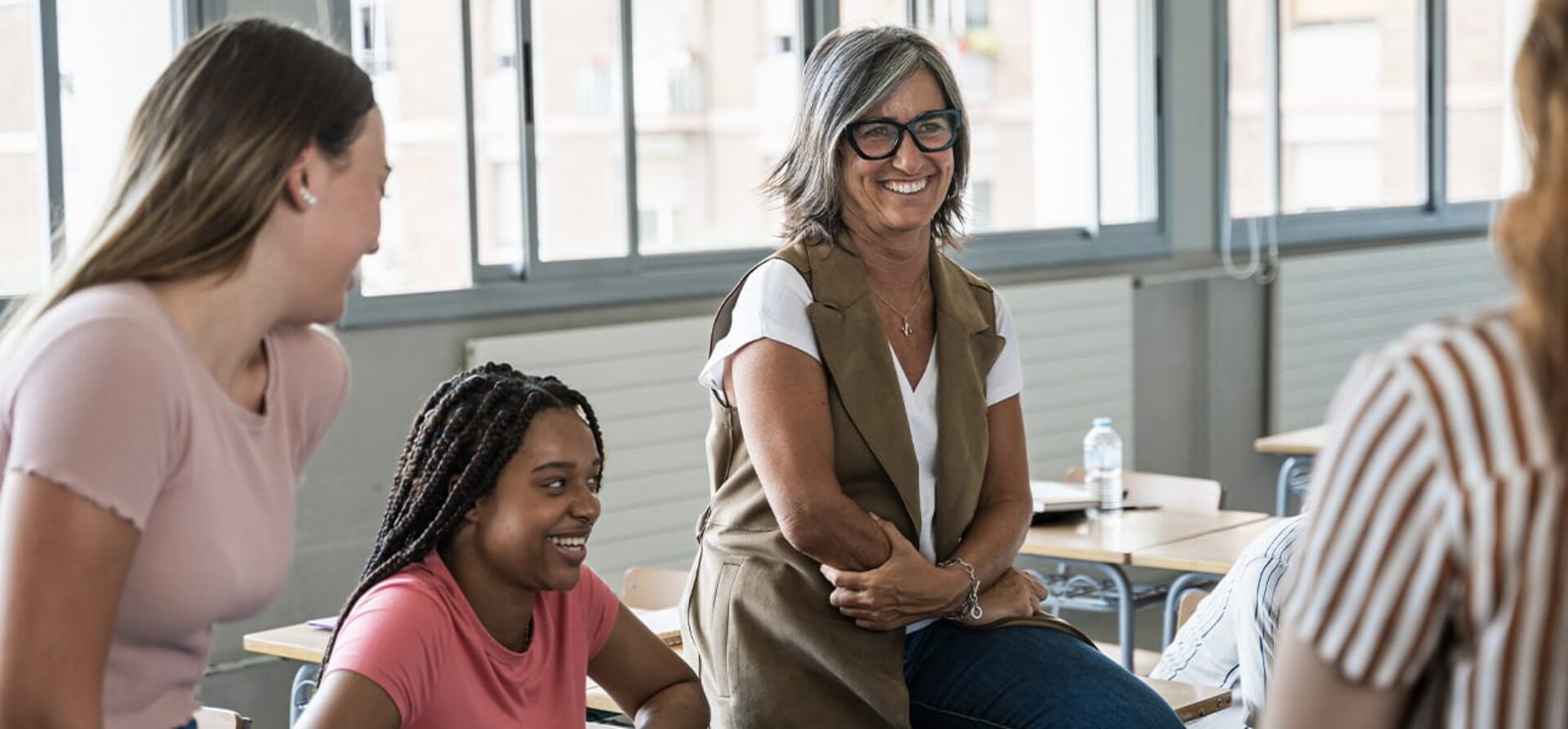 Group of female high students sit on school desks talking with female teacher
