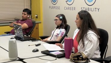 Three students sit at a long classroom table with laptops and notebooks